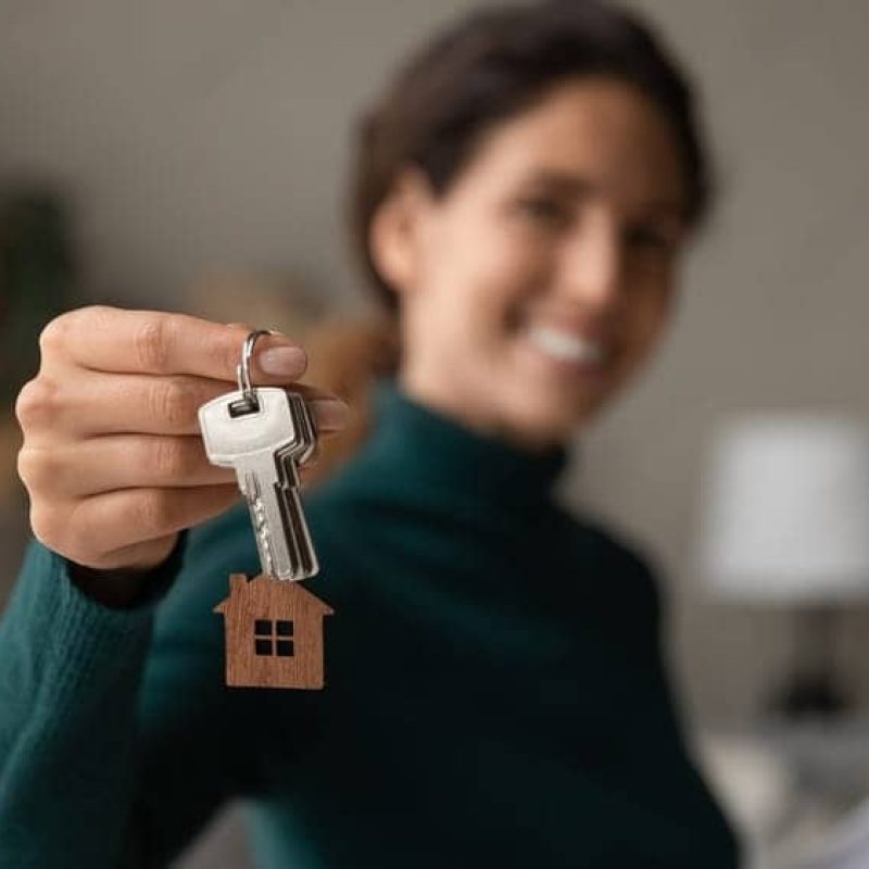 Close up focus on keys, smiling woman realtor selling apartment, offering to client, showing at camera, holding documents, contract, making purchasing